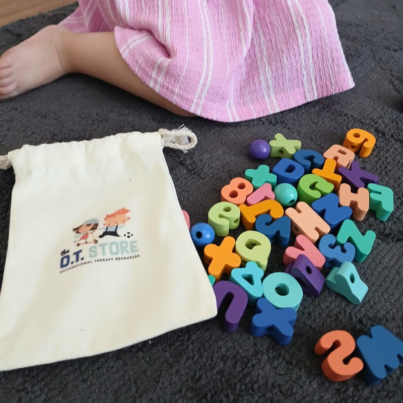 Colourful alphabet lacing beads scattered beside a branded drawstring bag on a dark carpet, next to a child wearing a pink striped dress.