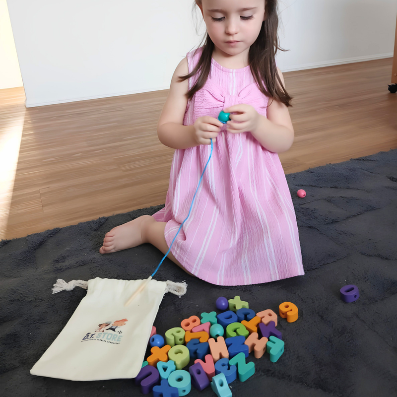 Young child sitting on a rug threading large colourful alphabet beads onto a blue string, with a drawstring bag beside them.
