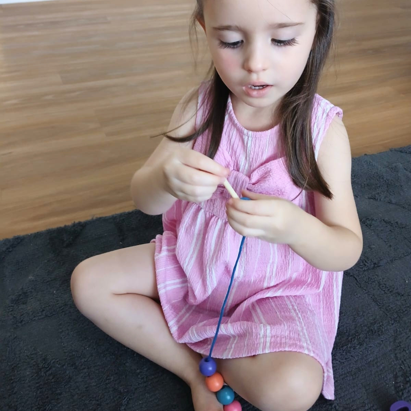 Young child in pink dress sitting on a rug threading colourful beads onto a blue string for fine motor play.
