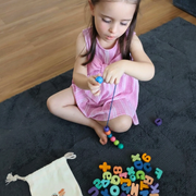 Young girl sitting cross-legged on a rug threading colourful alphabet lacing beads onto a blue string beside a cloth pouch.