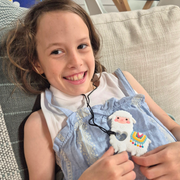 White and yellow llama chewable necklace on a black cord, held by a smiling child lounging on a sofa