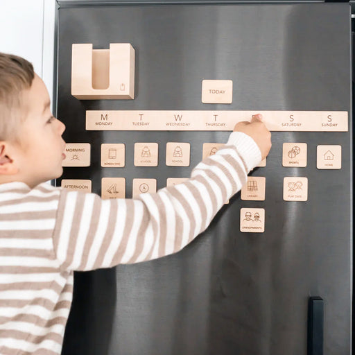 Child arranging wooden magnetic schedule pieces on a dark fridge, showing days of the week and daily activities in a home setting.