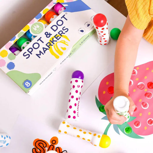 Child's hand using easy-grip spot and dot markers with colourful polka dots, next to their packaging and painted craft on paper.